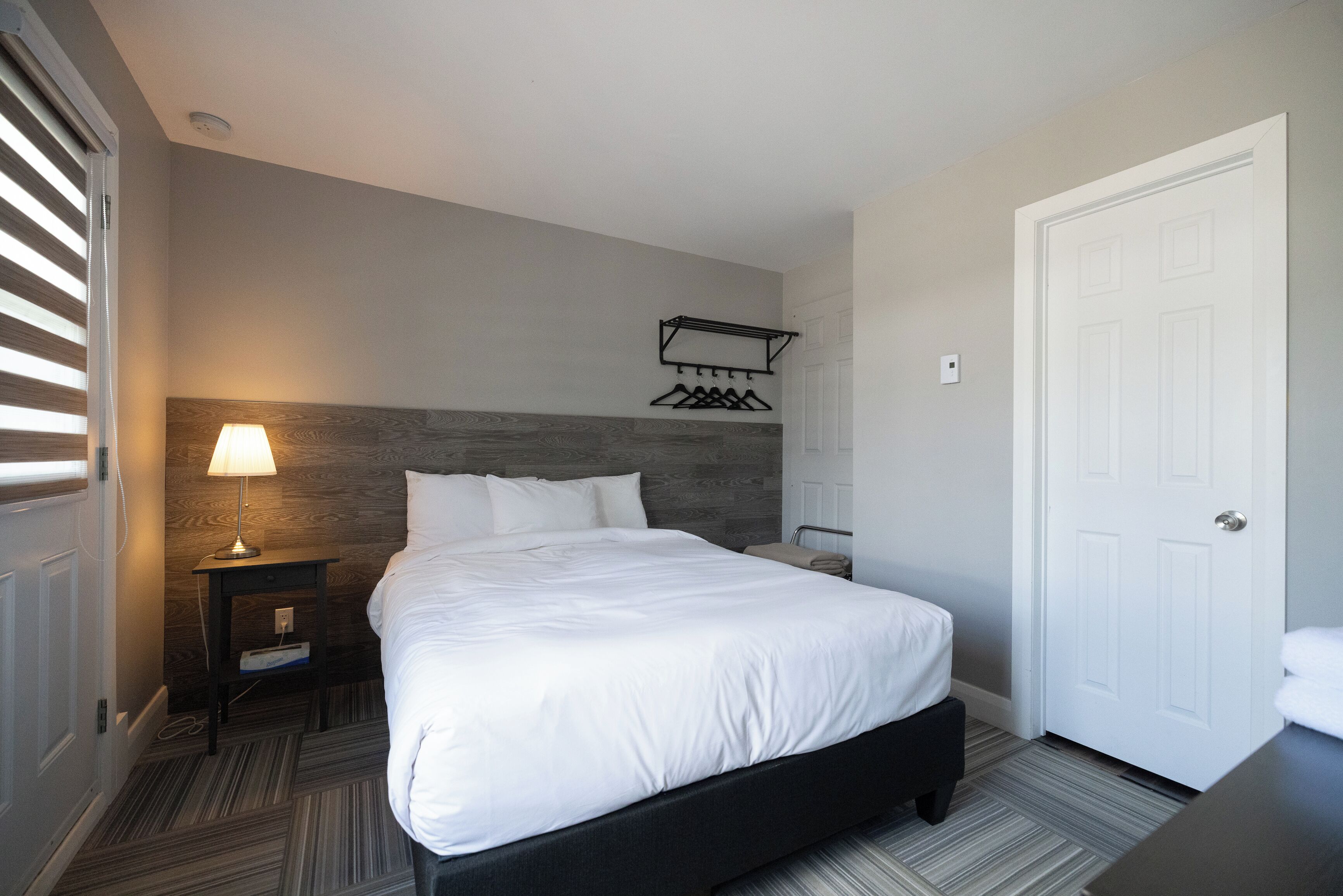 Ein Hotelzimmer mit einem Bett, einem Nachttisch mit Lampe, einem Kleiderständer und einer Tür.