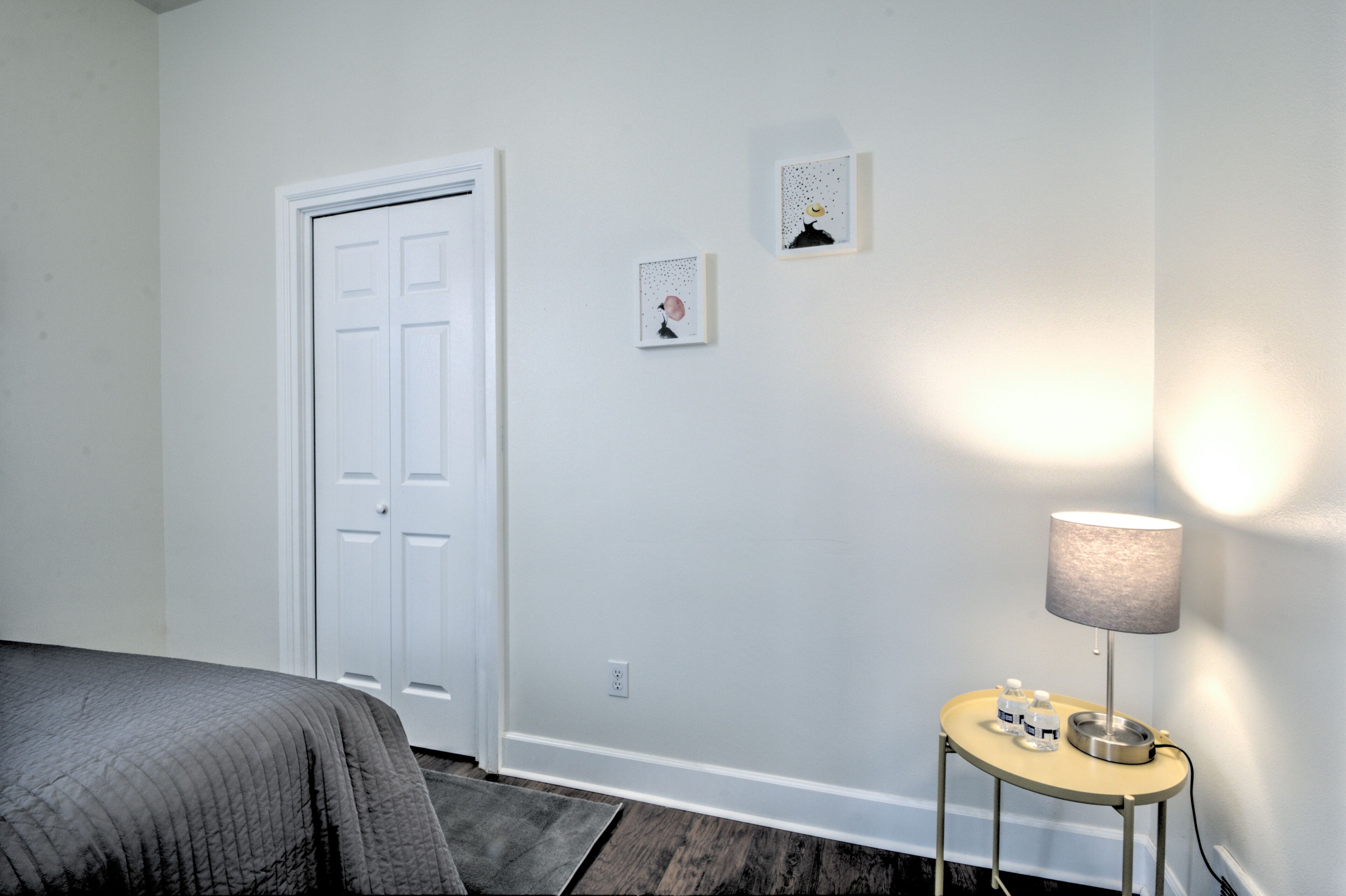 A bedroom with a bed, a door, a small table with a lamp, and two wall-mounted pictures.