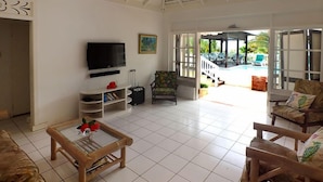 A spacious living area with a television, a small table, and a chair. There is a view of a pool and outdoor furniture. - Sea Spray 50 Steps to the Sea, Silver Sands, 3 BR (Duncans)