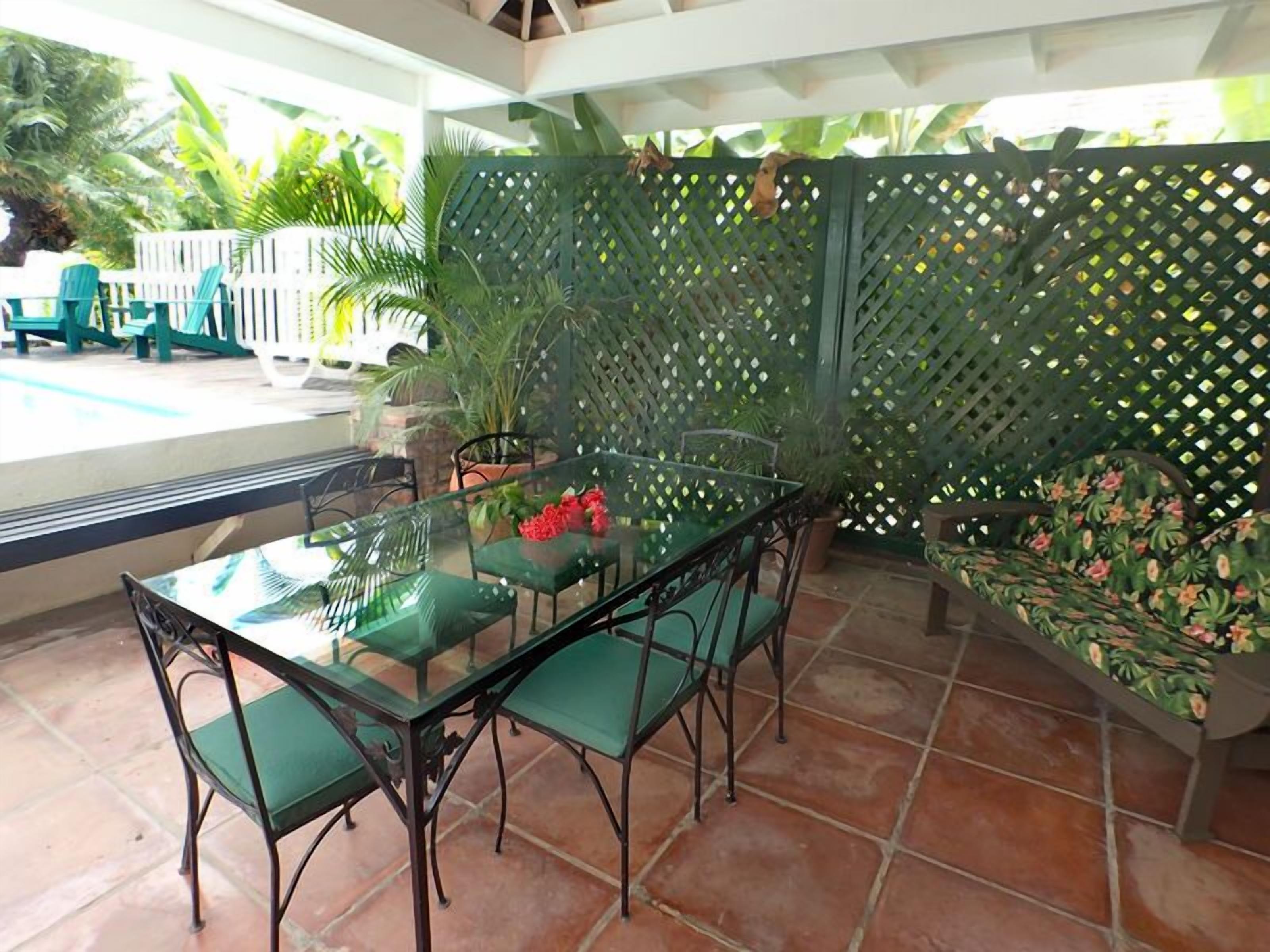 Une terrasse extérieure avec une table en verre et des chaises vertes, entourée d’une clôture en treillis et de plantes en pot.