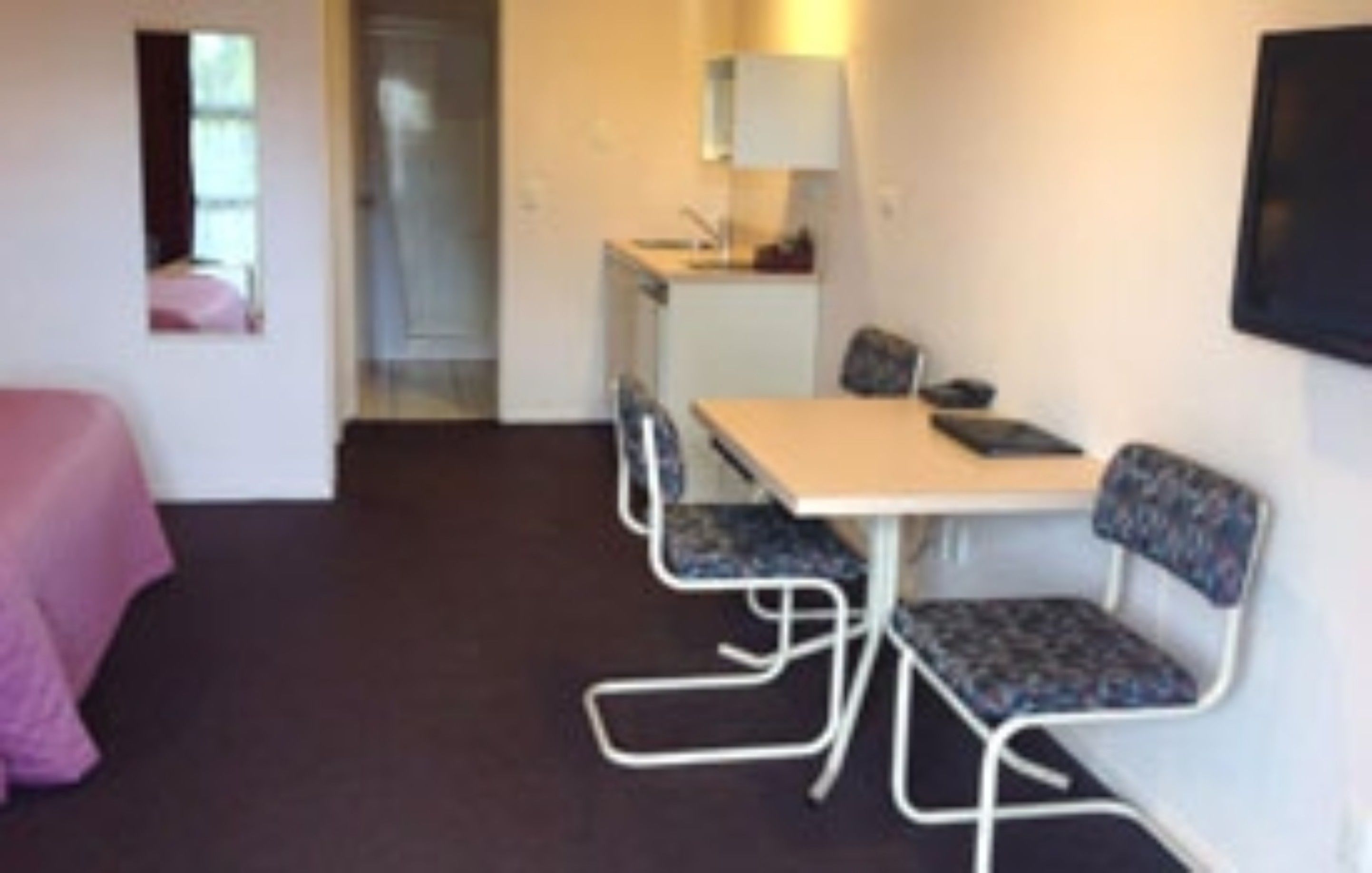 A compact room with a bed, a small kitchenette, a dining table with chairs, and a television mounted on the wall.