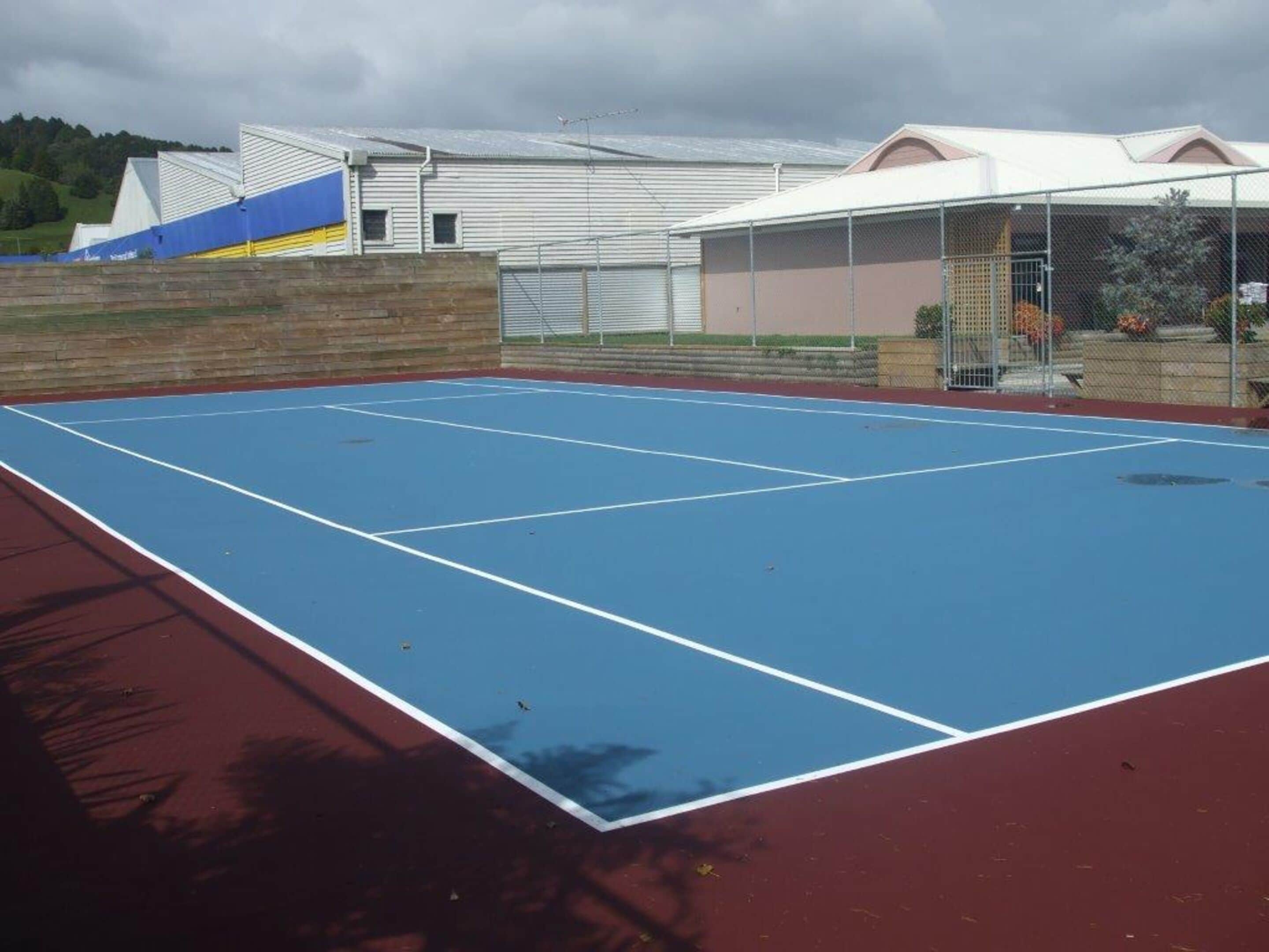 An outdoor tennis court with a red and blue surface, surrounded by a brick wall and a building with a white roof.