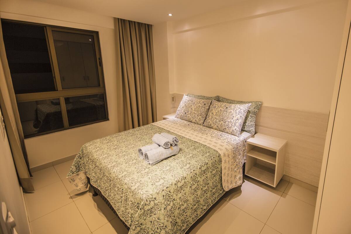 Una cama bien tendida con una colcha estampada con motivos florales y dos almohadas, una mesita de noche con una lámpara, y una ventana con cortinas.