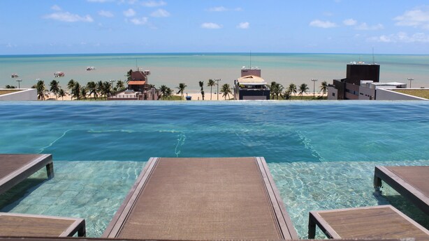 Una piscina infinita con agua cristalina, sillones de madera, vista a la playa y palmeras.