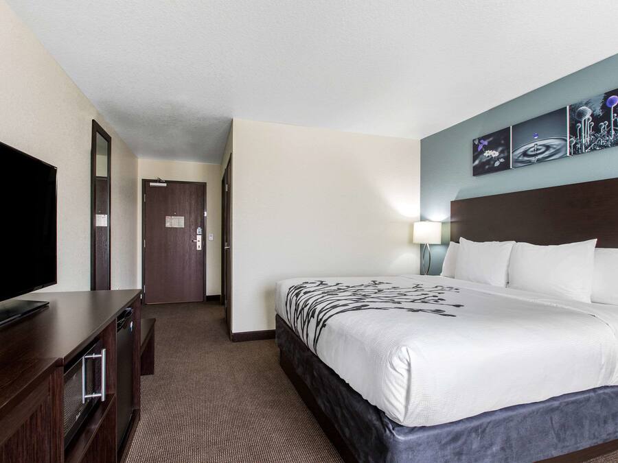Ein Hotelzimmer mit einem großen Bett, einem Flachbildfernseher, einer Holzkommode und einem an der Wand hängenden Kunstwerk.