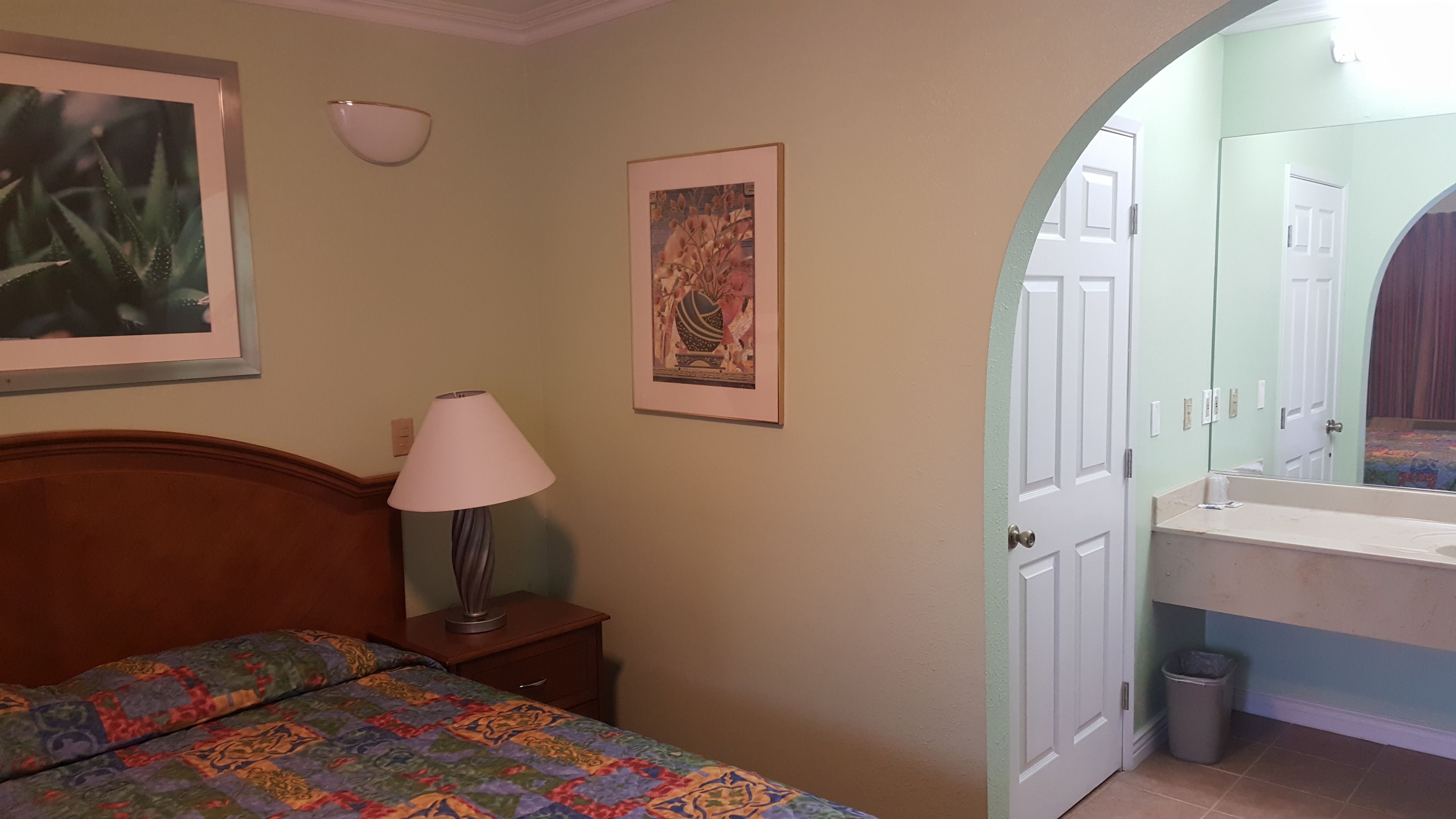 A bedroom with a bed, a bedside table with a lamp, a framed picture on the wall, and a bathroom with a sink and mirror.
