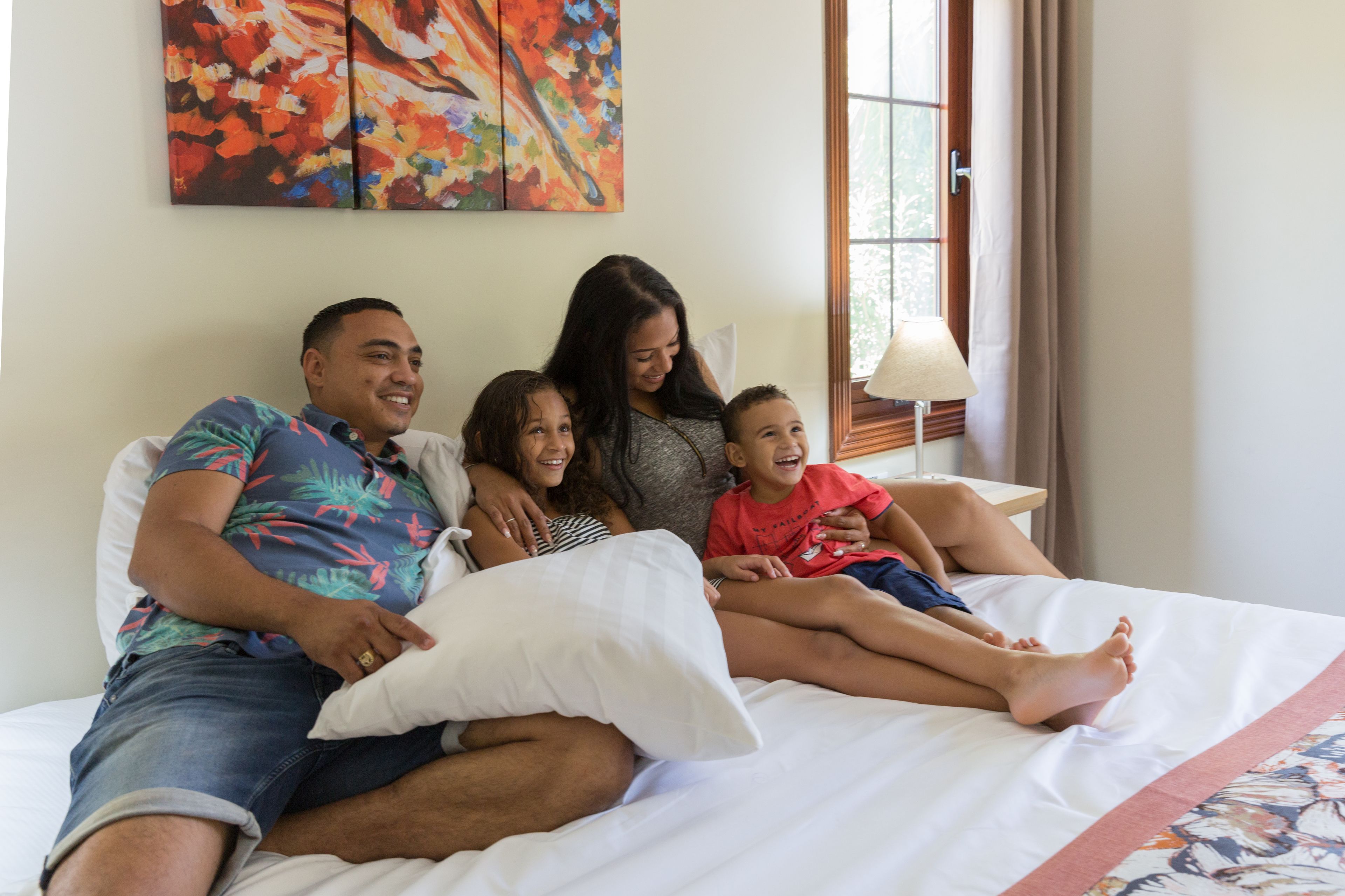 En familie på fire på en seng i et hotelværelse med et stort vindue, et billede på væggen og en sengetavlelampe.