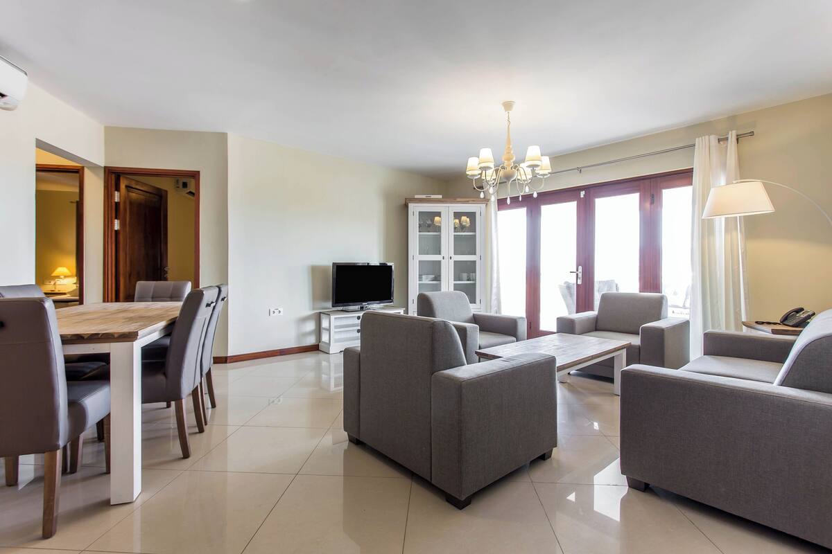A modern living room with a dining area, grey sofas, a TV, and a chandelier.