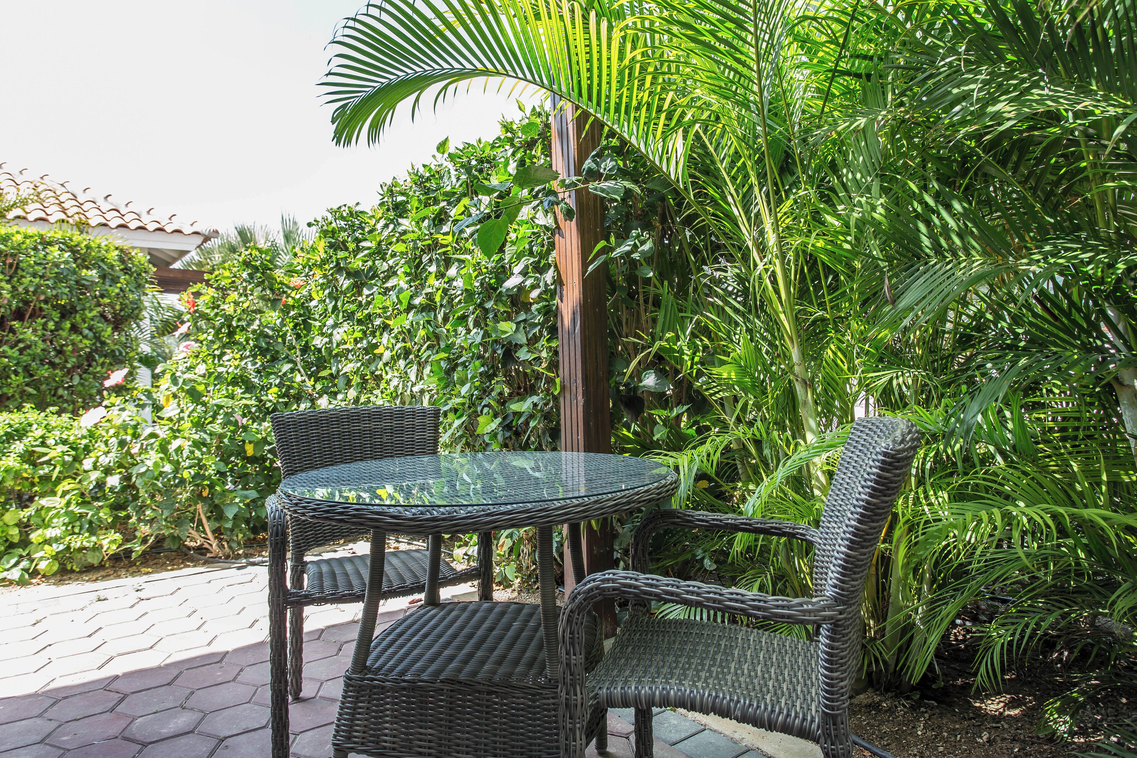 Une table à plateau en verre, entourée de chaises en osier, dans un coin de jardin verdoyant.