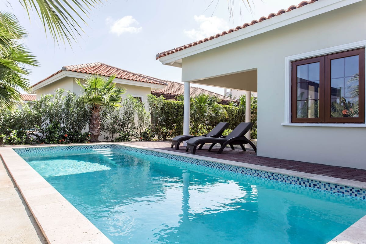 A swimming pool with lounge chairs, surrounded by greenery and a house with a tiled roof.