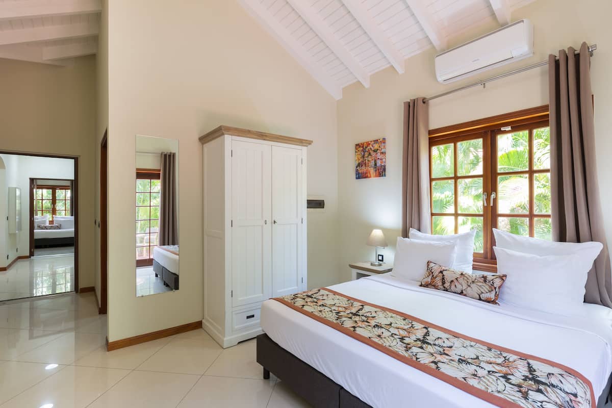 A bedroom with a large bed, a wardrobe, a mirror, a window with curtains, and an air conditioning unit.