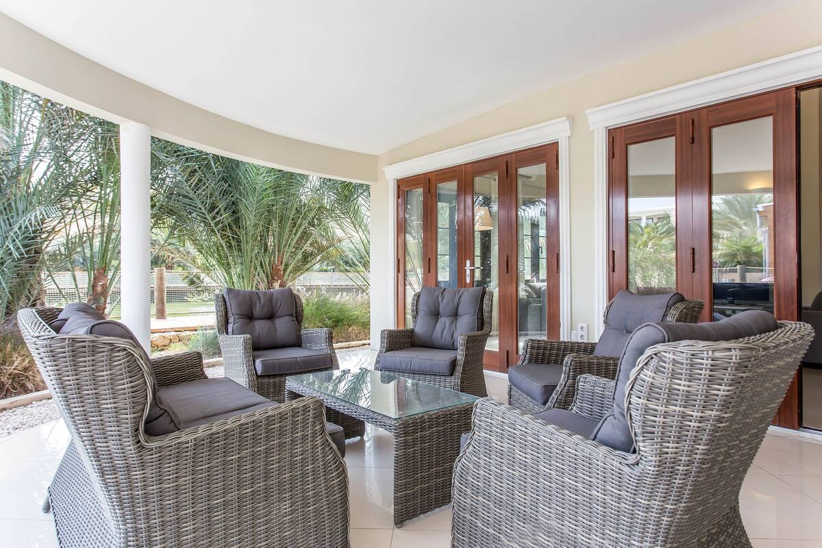 A covered outdoor seating area with wicker furniture and glass-top tables, surrounded by glass doors and palm trees.