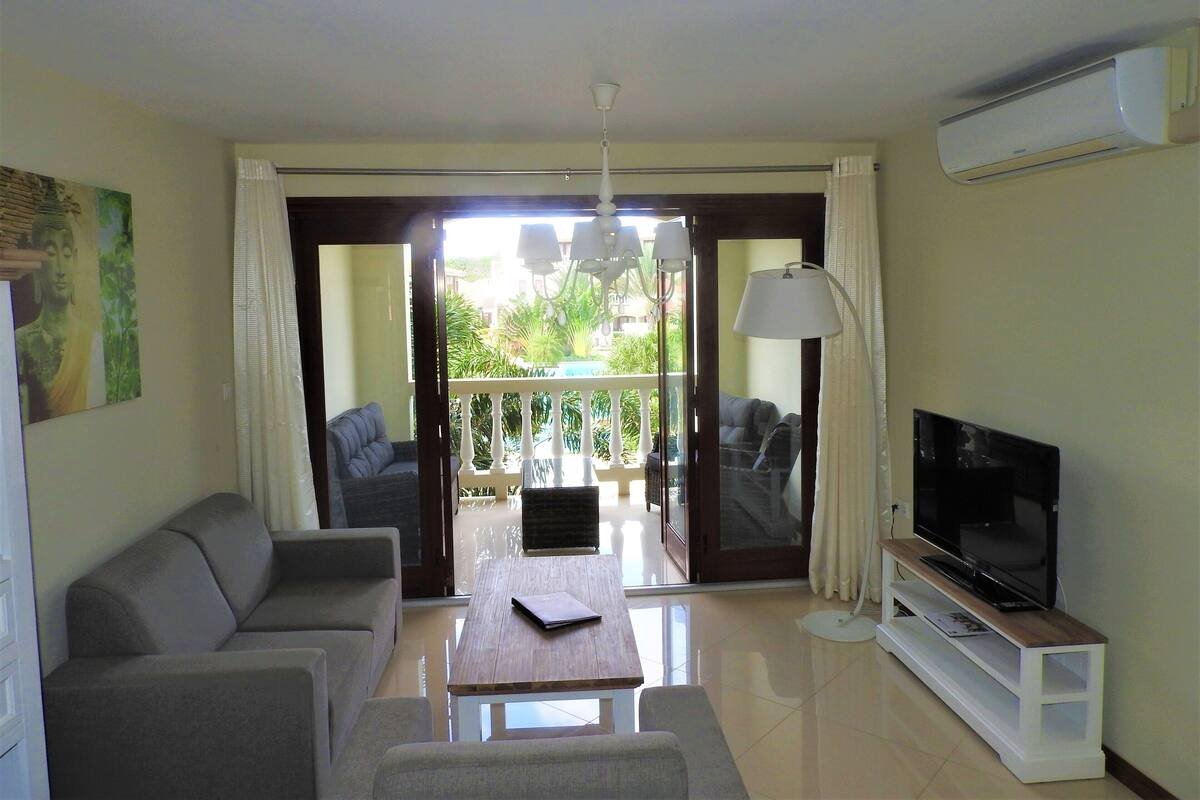 A living room with a sofa, a coffee table, a TV on a stand, and a balcony with a view.