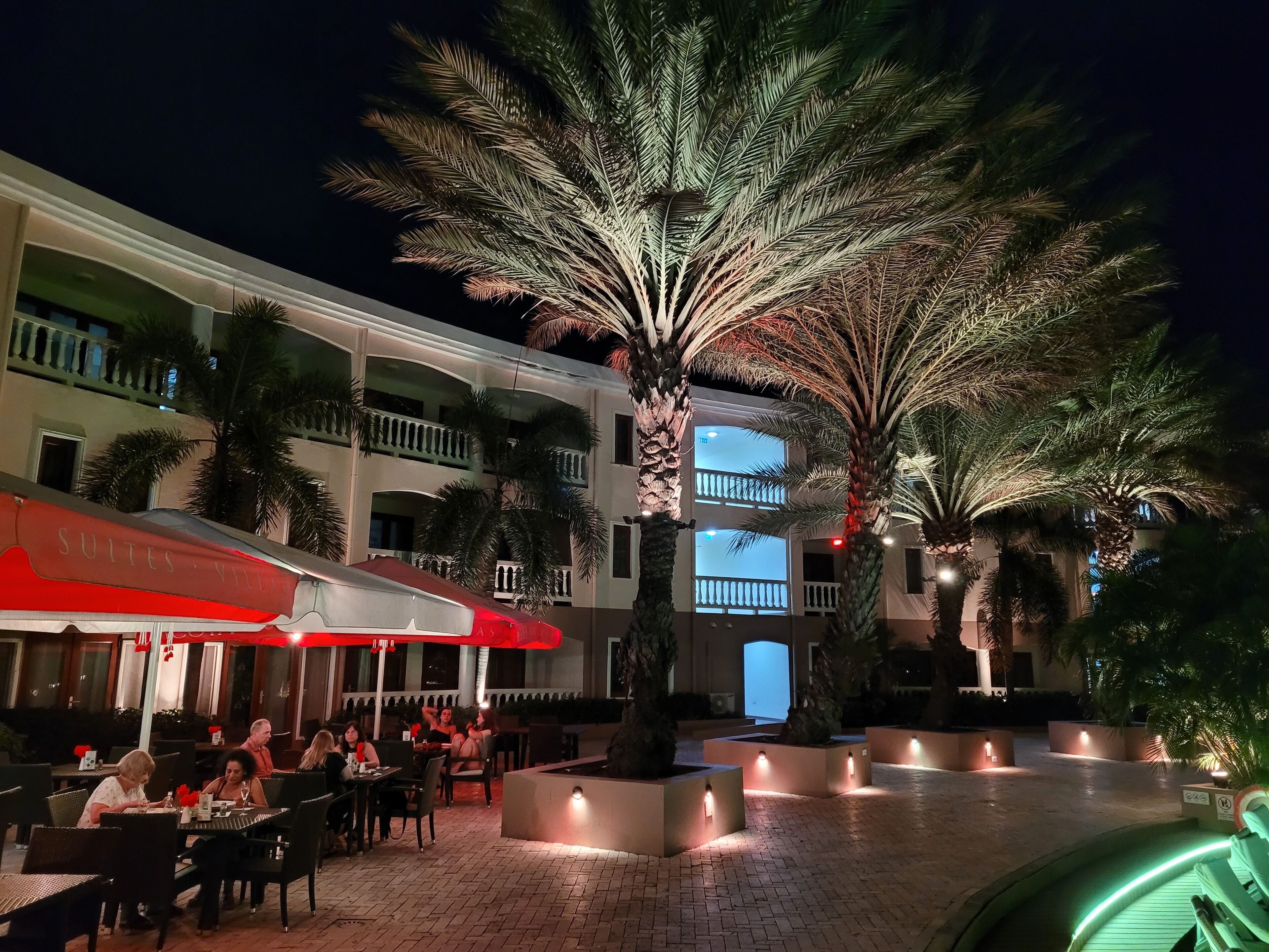 Patio de hotel con mesas al aire libre, palmeras y un edificio de varios pisos con balcones.