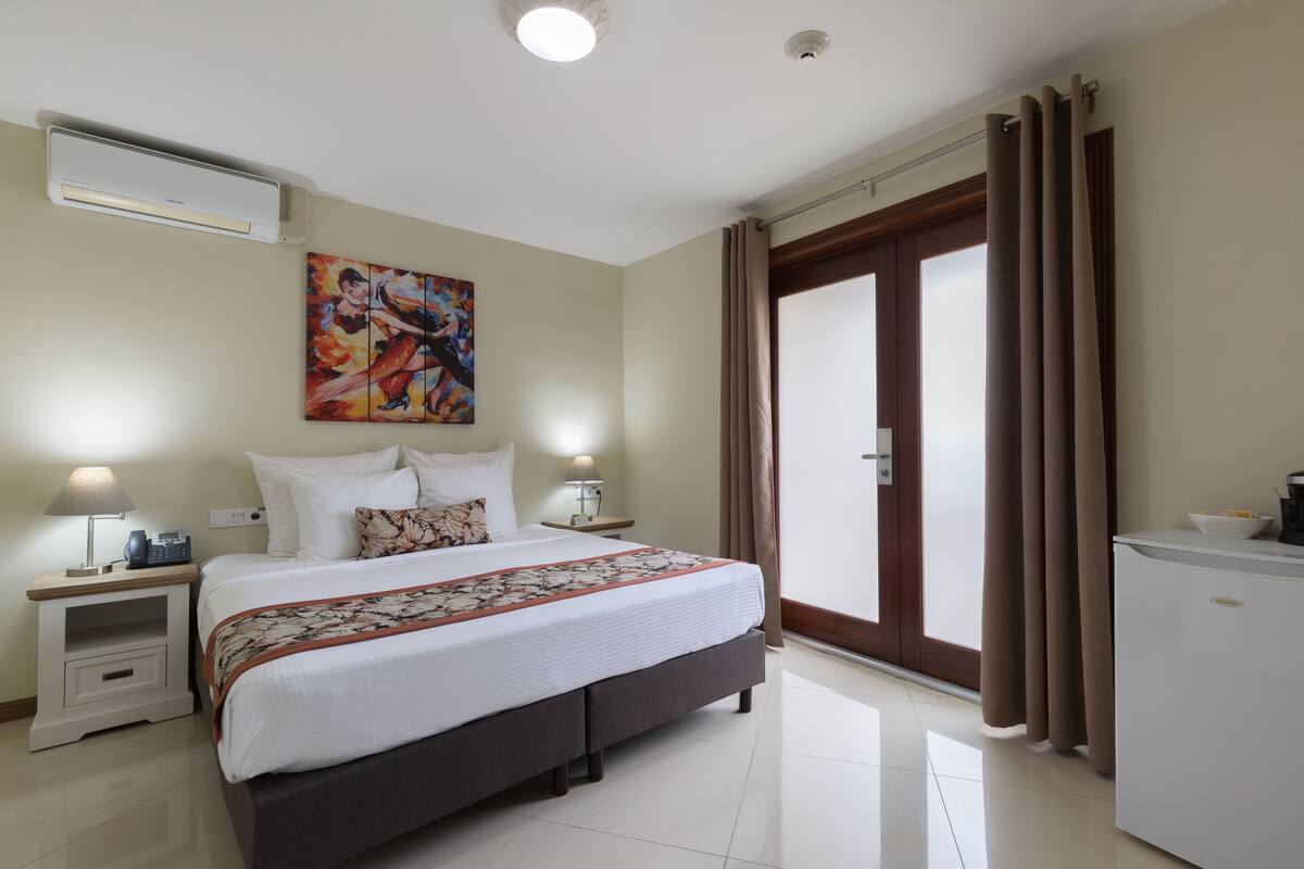 A hotel room with a large bed, a nightstand with a lamp, a painting on the wall, and a door with curtains.