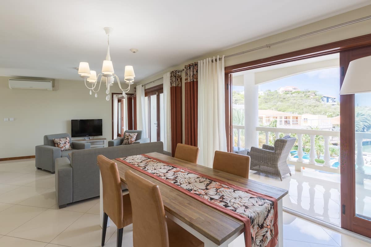 A modern living room with a dining area, a flat-screen TV, and a balcony with a view of a hill and buildings.