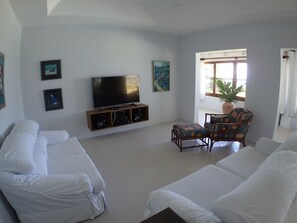 Una sala de estar con un televisor de pantalla plana, dos sofás blancos, un sillón estampado y una planta en maceta junto a una ventana.