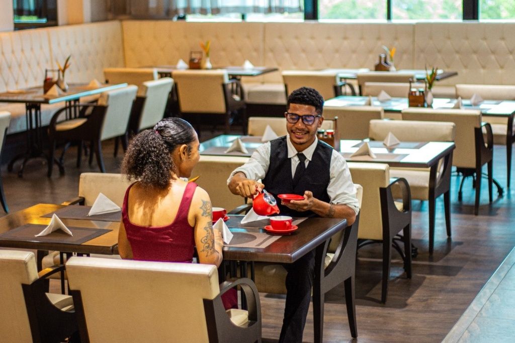 A restaurant interior with multiple tables and chairs, a waiter serving a customer.