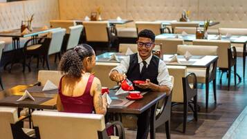 A restaurant interior with multiple tables and chairs, a waiter serving a customer.