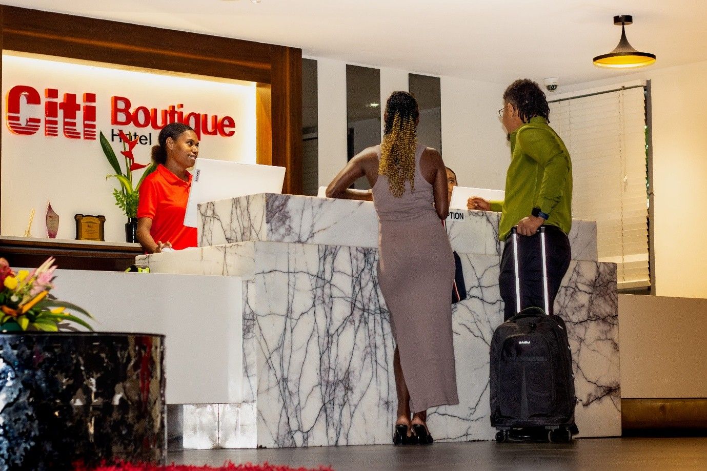 A hotel reception desk with a marble design, a receptionist, and guests standing in front of it.