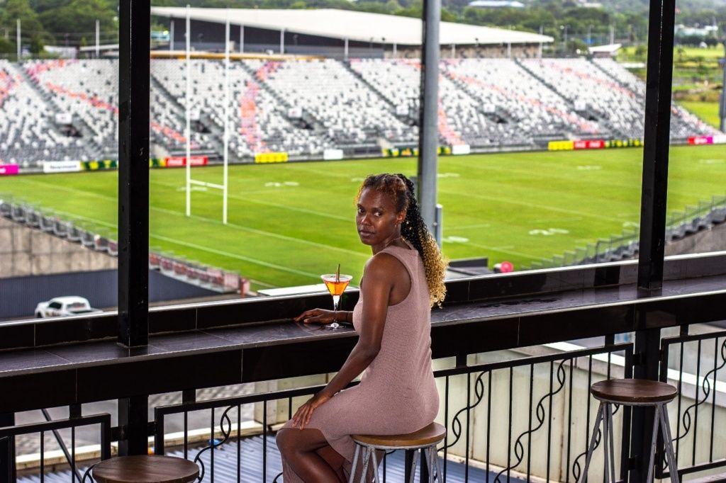 A person seated on a stool by a window overlooking a stadium with empty stands.