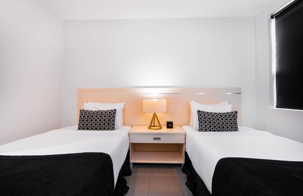 A hotel room with two beds, a bedside table with a lamp, and a TV mounted on the wall.