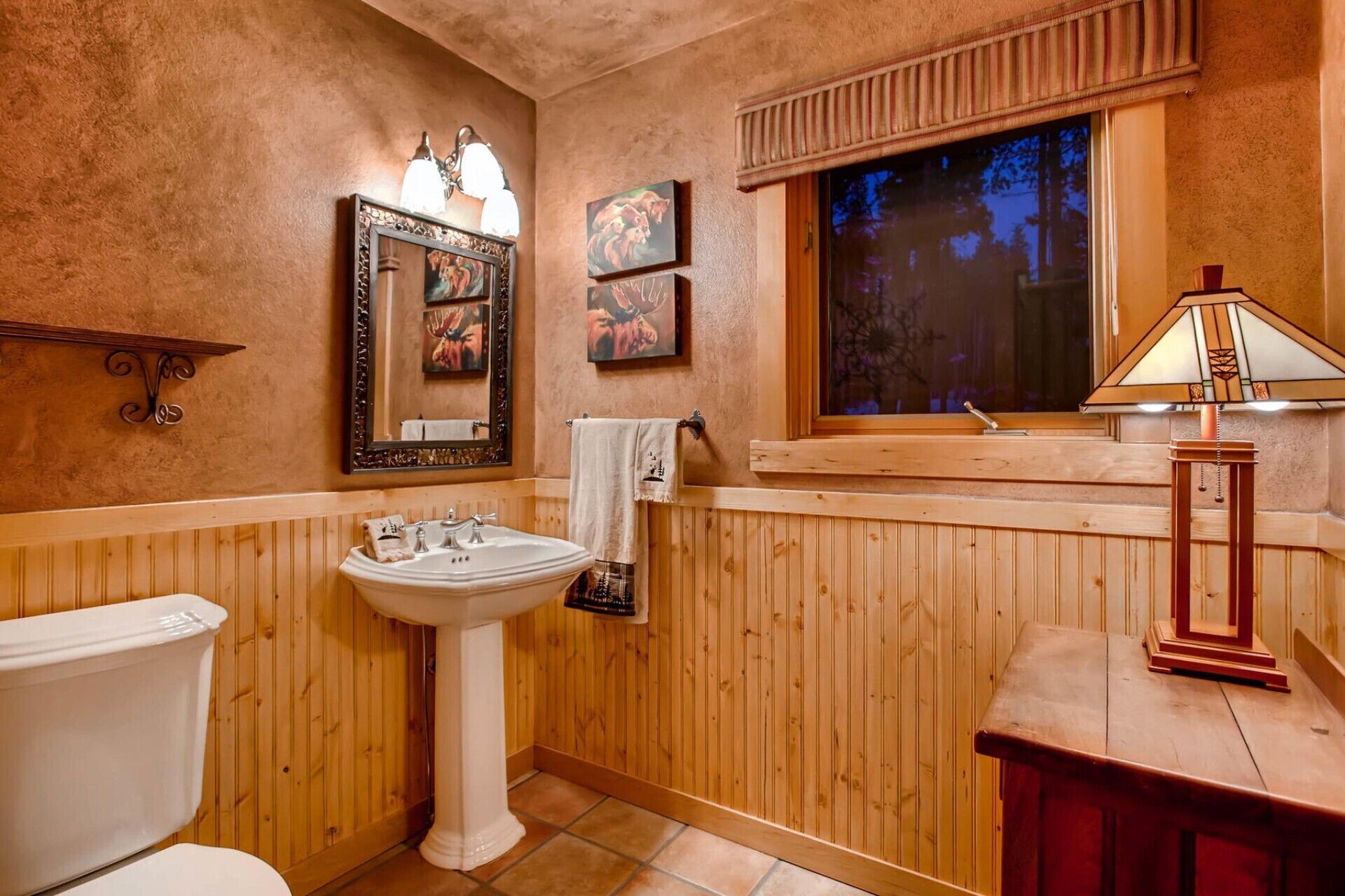 Un baño con paneles de madera, un lavamanos pedestal, un inodoro, un tocador con espejo y una ventana con vista a árboles.
