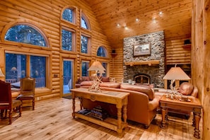A log cabin interior with a stone fireplace, leather sofas, and a wooden dining table.