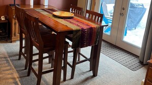 Un comedor con una mesa de madera, sillas y un mantel corrido estampado. Se observan obras de arte enmarcadas en la pared, una lámpara y vistas a través de un ventanal.