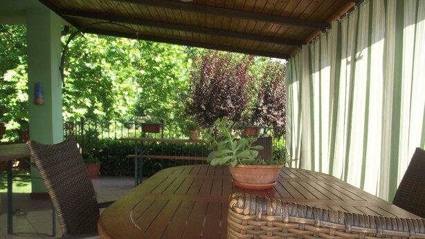 Un porche cubierto con una mesa de madera y sillas de mimbre, rodeado de plantas en macetas y una valla verde.