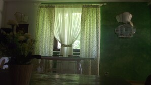 Una habitación con pared verde, ventana con cortinas florales, jaula de pájaros en la pared y planta en maceta.
