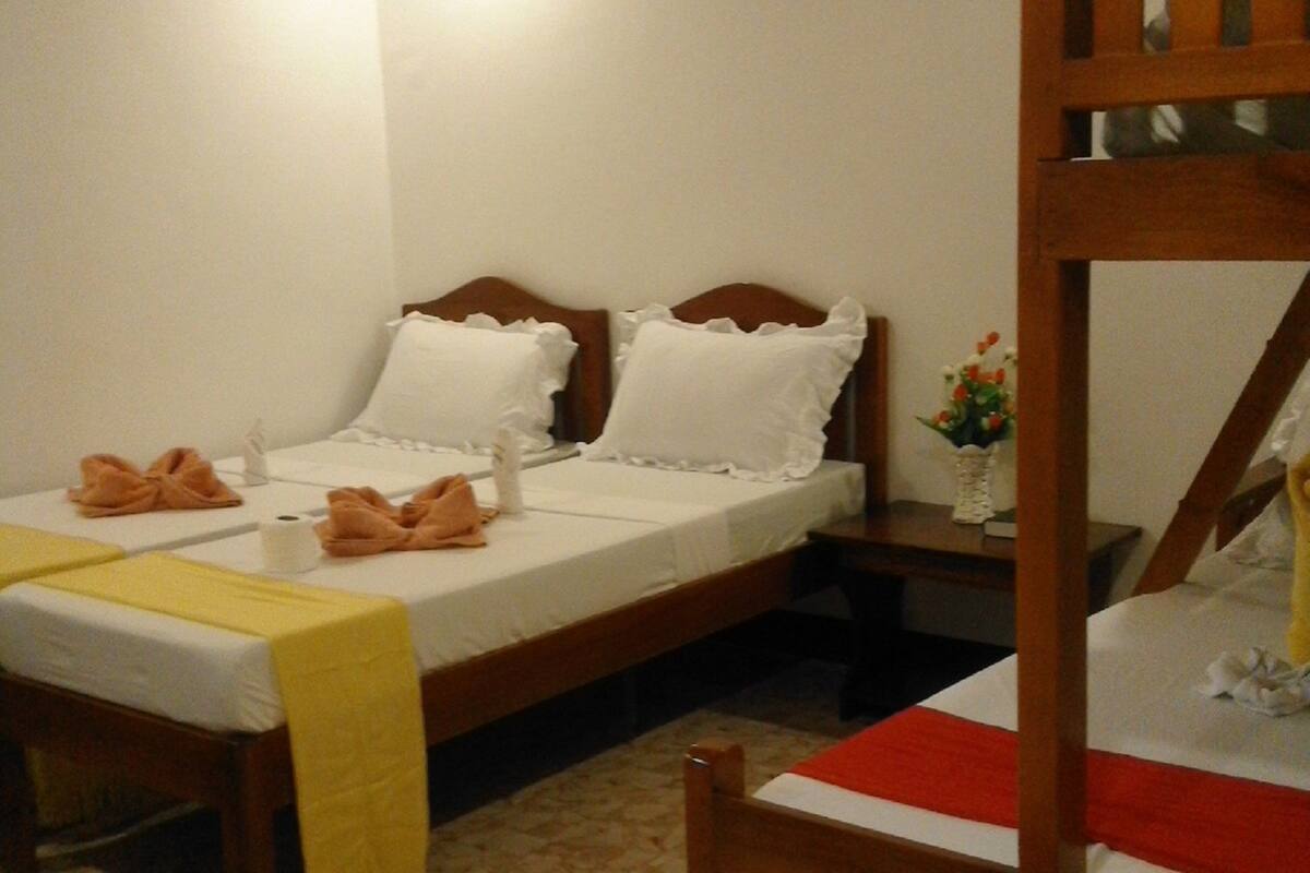 A room with two beds, one with a wooden headboard and the other with a white headboard. There is a small table with a vase of flowers and a yellow towel on the bed.
