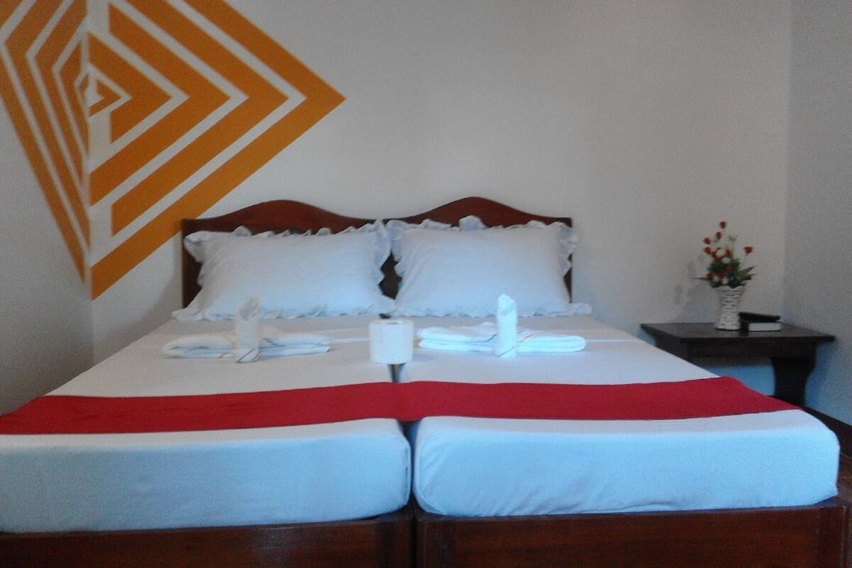 A double bed with white and red bedding, two pillows, a white vase with flowers, and a geometric wall design.