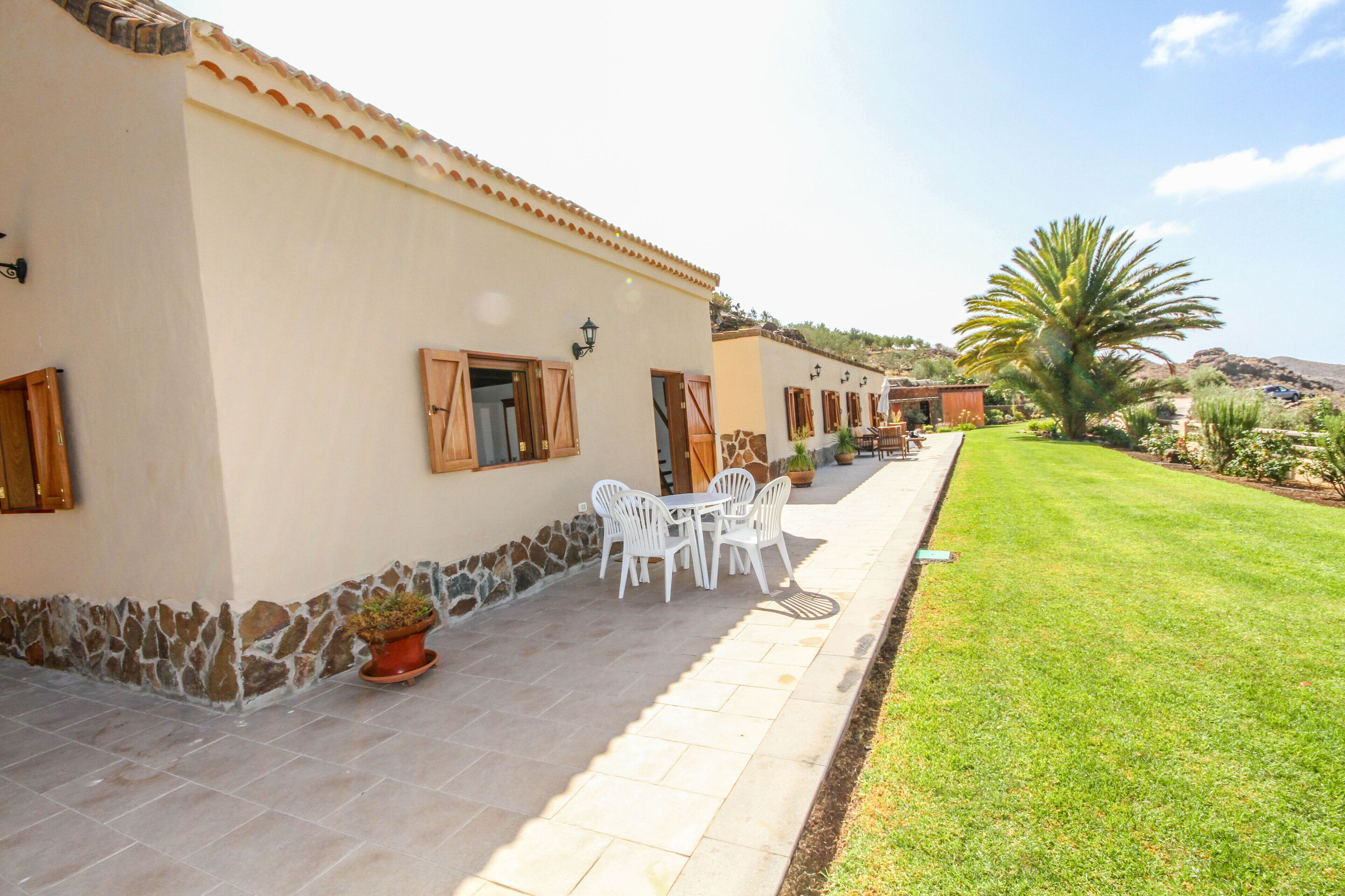 A building with a stone wall, a patio with white chairs and a table, and a well-maintained lawn.