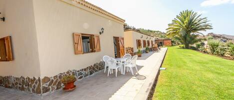 A building with a stone wall, a patio with white chairs and a table, and a well-maintained lawn.