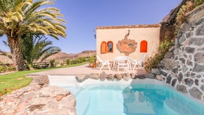 Zona de piscina con muro de piedra, una palmera y un edificio con ventanas de arco y un cartel.
