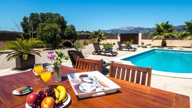 Ein Essbereich am Pool mit Tisch, gedeckt für ein Frühstück, darunter Früchte und ein Getränk, mit Blick auf einen Pool und Berge im Hintergrund.