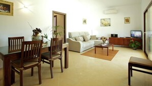 Ein Wohnzimmer mit einer Couch, Sesseln, einem Couchtisch und einem Fernseher. An den Wänden befinden sich Pflanzen und Kunstwerke.