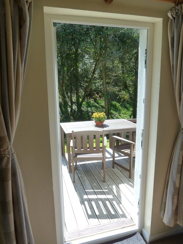 An open sliding door leading to an outdoor deck with a wooden table and chairs, a potted plant, and a view of a wooded area.