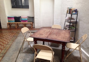 Una habitación con una mesa de madera rodeada por cuatro sillas, una estantería con libros, un banco con cojines y una alfombra estampada.