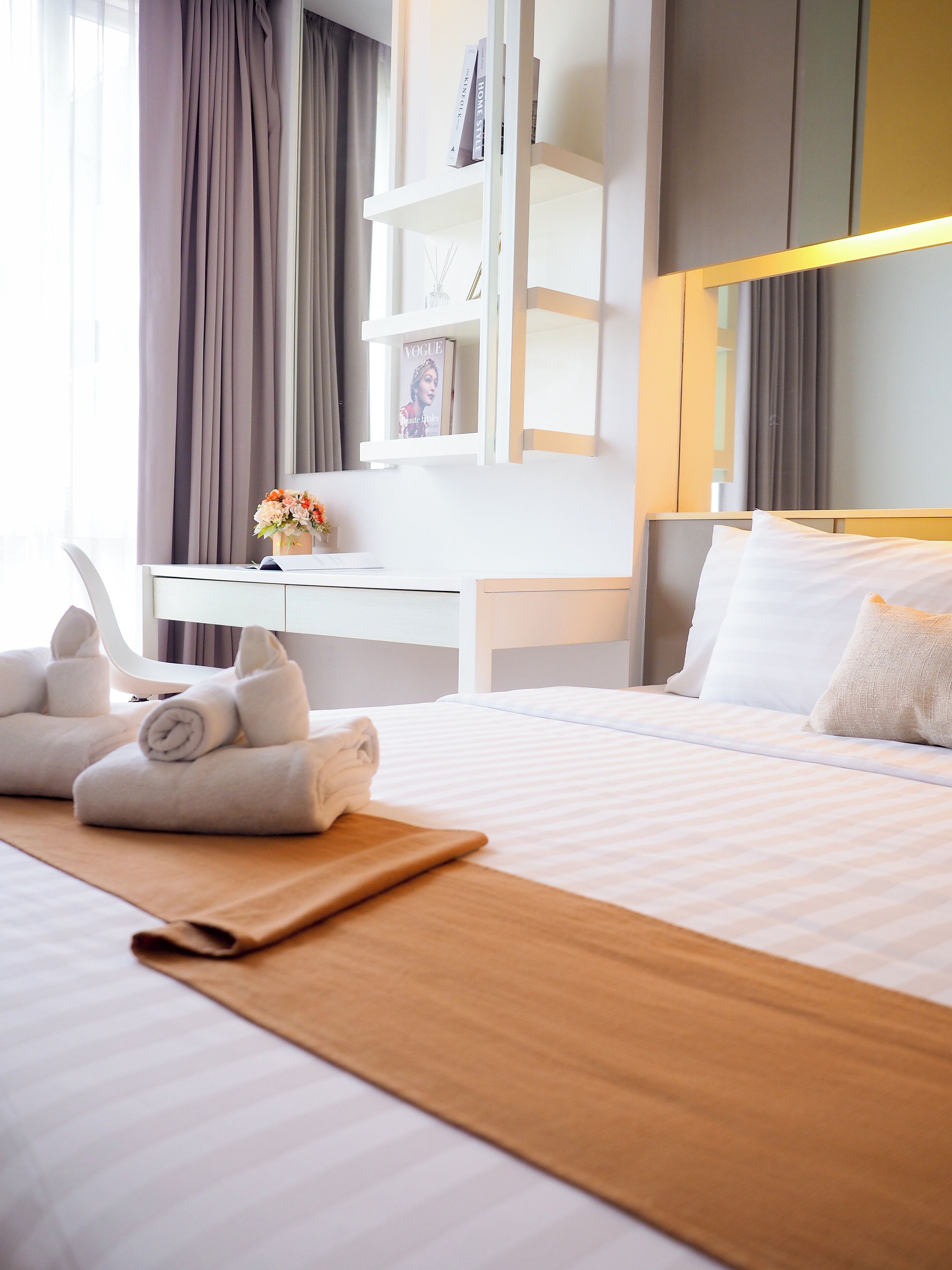 A hotel room with a neatly made bed, a desk with a chair, a bookshelf, and a large mirror.