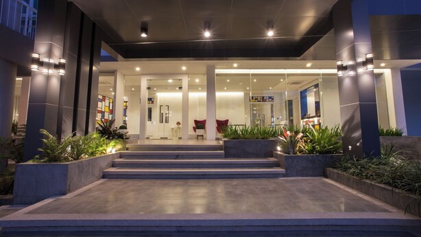 A modern hotel entrance with a glass facade, steps leading up to the entrance, and indoor plants. - The Kannas Hotel (Chiang Mai)