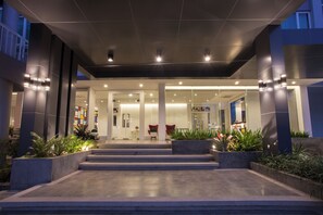 A modern hotel entrance with a glass facade, steps leading up to the entrance, and indoor plants. - The Kannas Hotel (Chiang Mai)