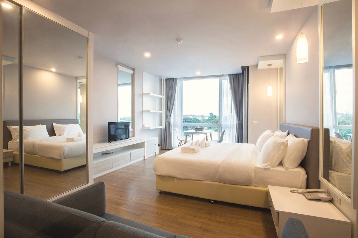 A modern hotel room with a large bed, a TV, a desk with a phone, and a balcony with a view.