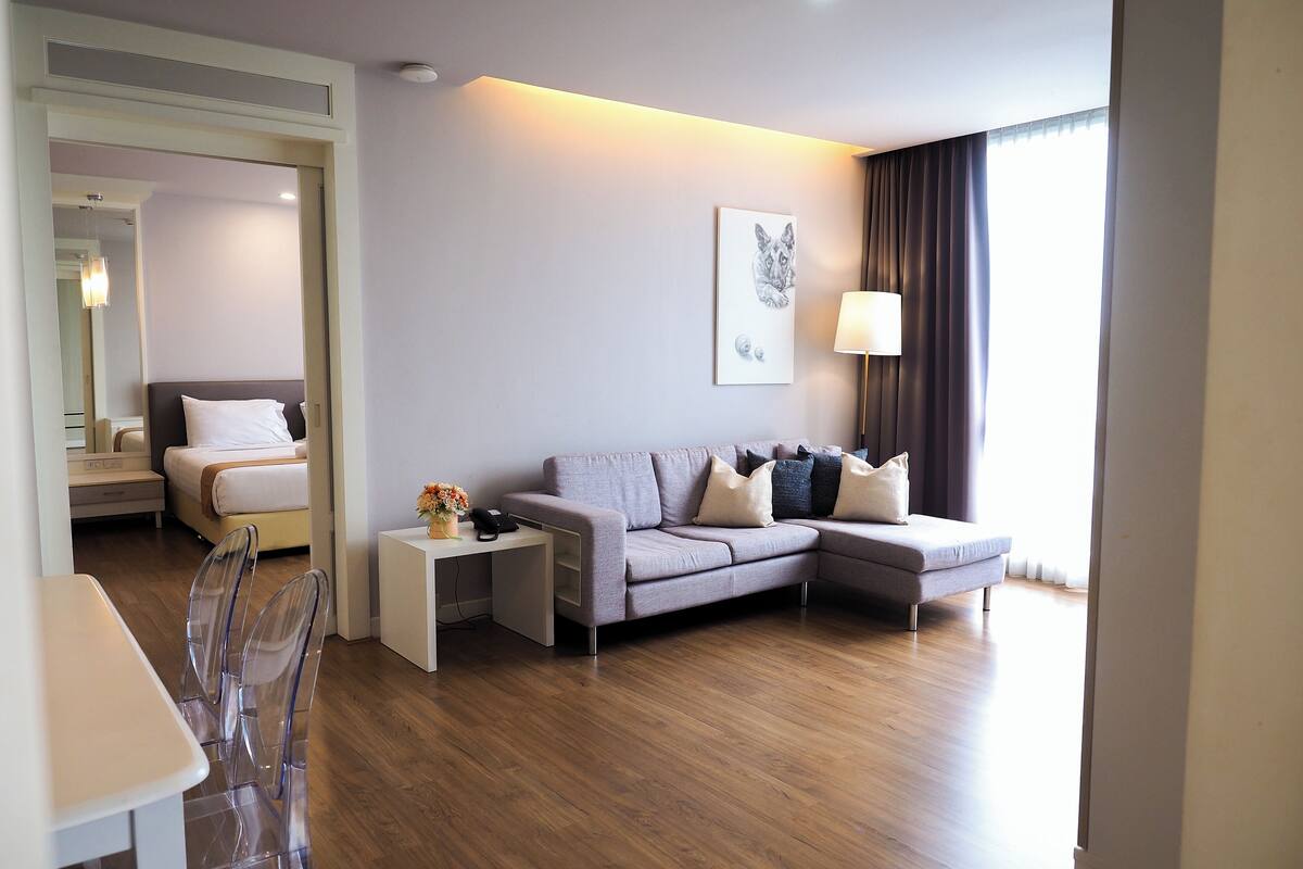 A modern living room with a grey sofa, a small side table, a clear chair, and a dining table.