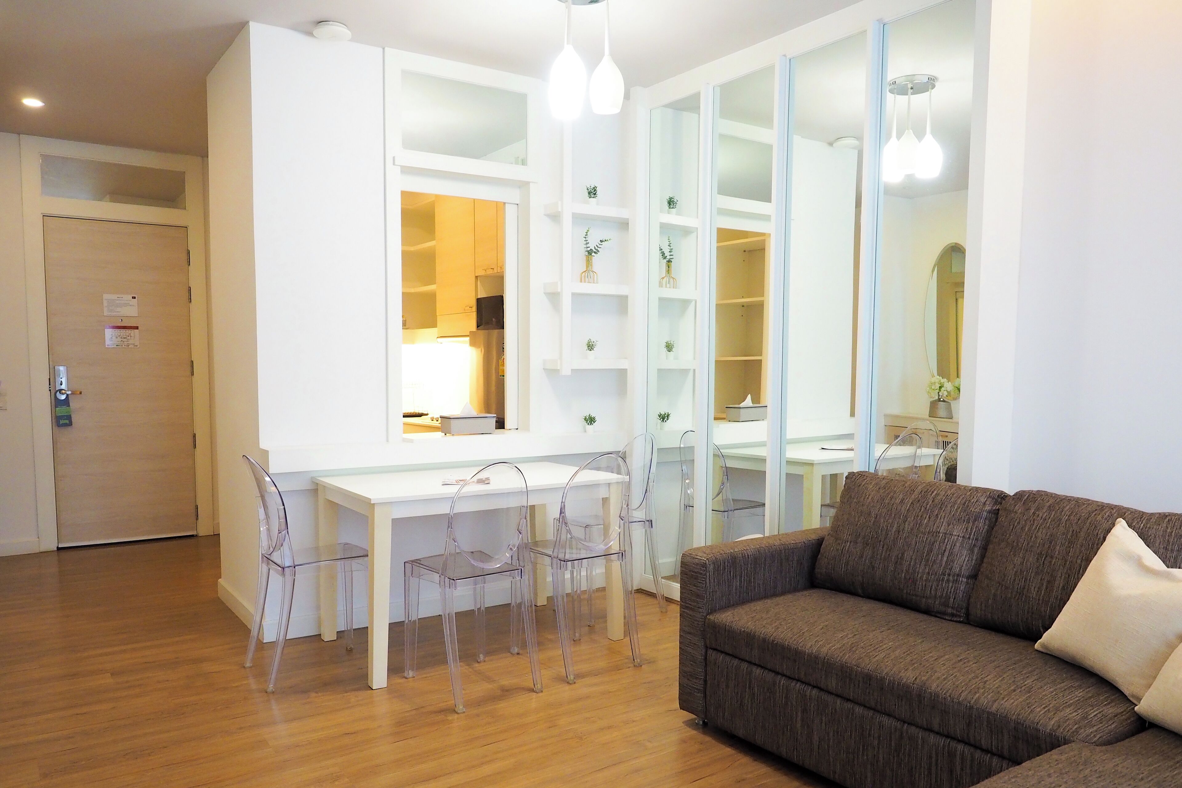 Ein modernes Wohnzimmer mit Holzboden, einem weißen Schreibtisch, transparenten Stühlen, einem braunen Sofa und einem großen Spiegel.