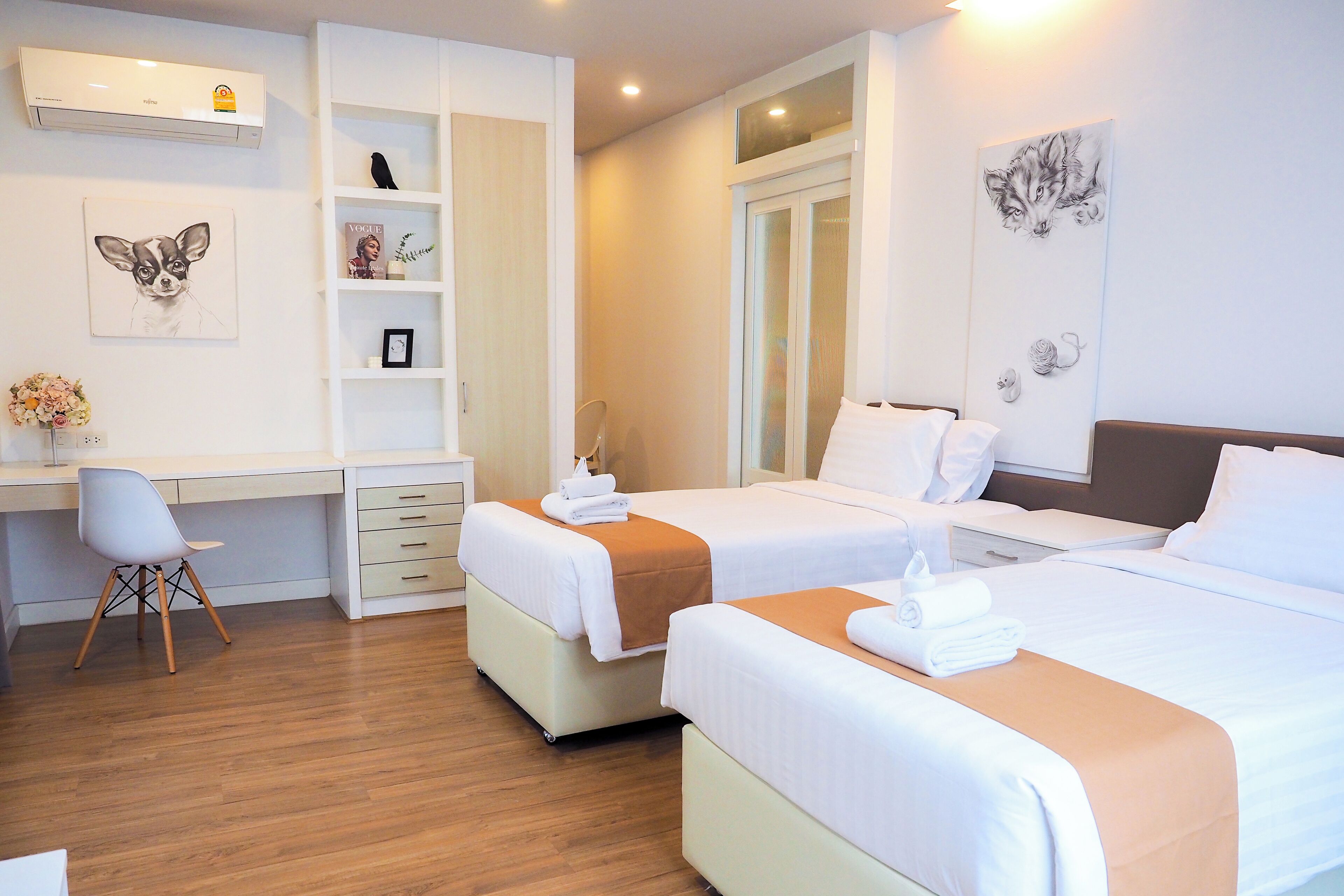 A hotel room with two beds, a desk, and a chair. There is a bookshelf and a wall-mounted air conditioner.