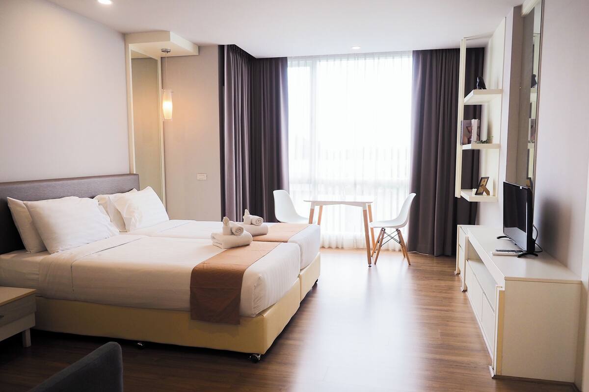 A modern hotel room with a large bed, a desk, a chair, and a balcony with curtains.