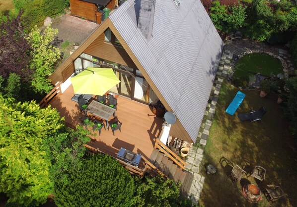 A triangular wooden house with a deck, outdoor seating, and a swimming pool.