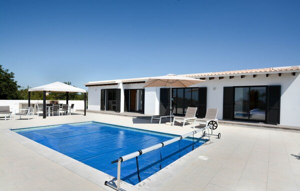 A modern outdoor pool area with a clear blue pool, sun loungers, a covered seating area, and a building with large windows and shutters. - Casa da Vinha - Carvoeiro - Pool villa for 6 persons with sea view on pur nature (Carvoeiro)